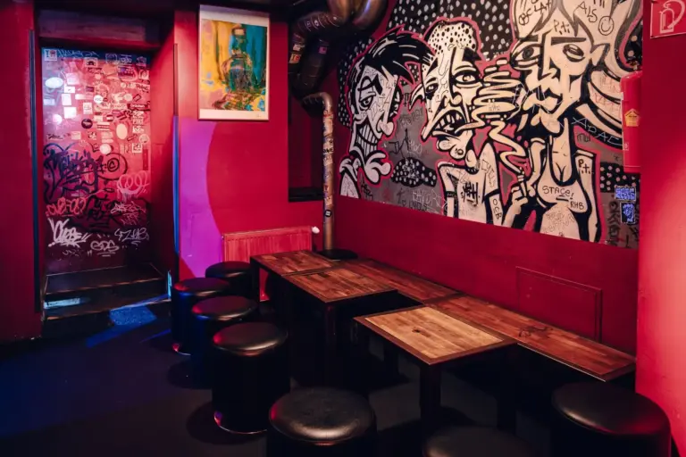 Eine schummrig beleuchtete Bar mit roten Wänden, schwarzen Hockern und Holztischen. An den Wänden befinden sich Graffiti-Kunstwerke mit ausdrucksstarken Gesichtern und eine Collage aus Aufklebern neben der Tür. Rosa und blaues Umgebungslicht erfüllt den Raum.