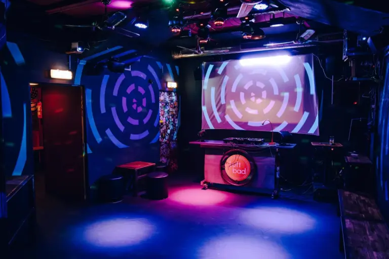 Ein farbenfroher Nachtclub mit kreisförmigen Lichtmustern an den Wänden, einem DJ-Pult in der Mitte und lila, blauen und rosa Lichtern, die die leere Tanzfläche beleuchten.