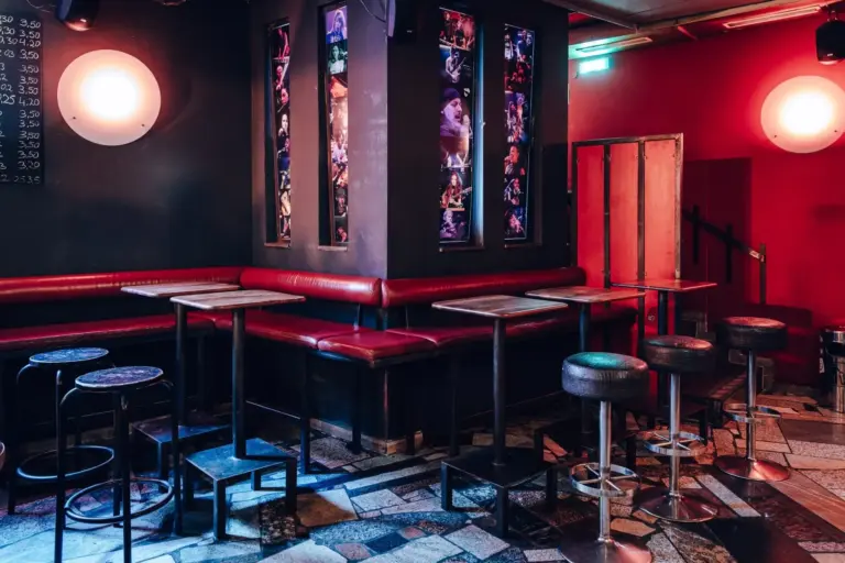 Eine schummrig beleuchtete Bar mit roten Wänden und Sitzbänken, leeren Tischen und Hockern und vertikalen Fototafeln von Musikern an der Eckwand. Der Boden ist mit uneinheitlichen Fliesen gemustert.