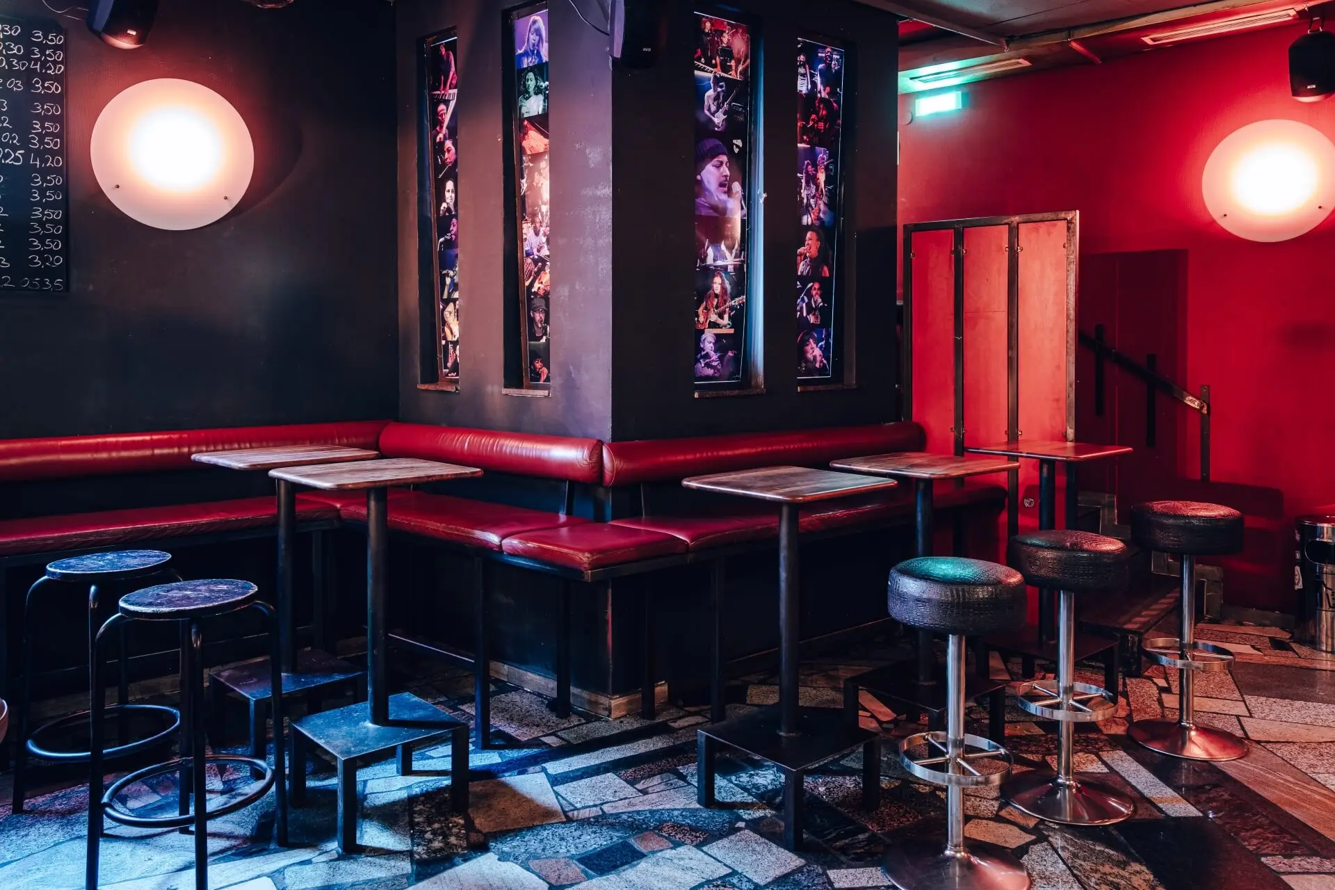 Eine schummrig beleuchtete Bar mit roten Wänden und Sitzbänken, leeren Tischen und Hockern und vertikalen Fototafeln von Musikern an der Eckwand. Der Boden ist mit uneinheitlichen Fliesen gemustert.
