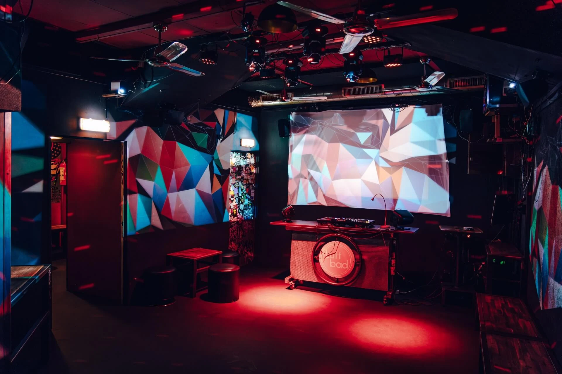 Ein schwach beleuchteter Nachtclub mit roter Beleuchtung, einer DJ-Kabine, geometrisch gemusterter Wandkunst, leeren Hockern und einem großen Bildschirm, auf dem bunte abstrakte Formen zu sehen sind.
