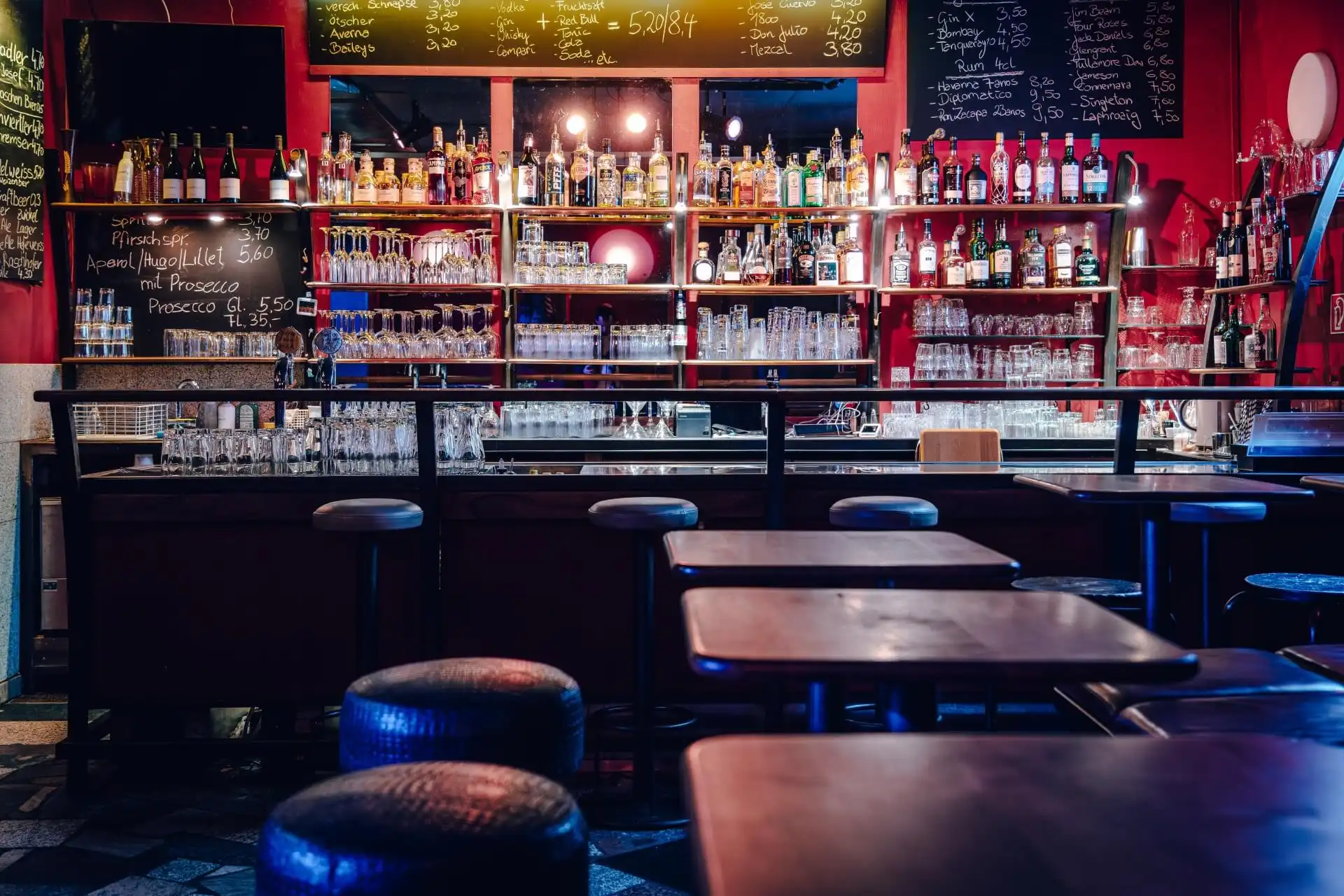 Eine schummrig beleuchtete Bar mit leeren Hockern und Tischen im Vordergrund. Im Hintergrund befinden sich Regale mit Schnapsflaschen, Gläsern und einer Speisekarte, die auf Kreidetafeln über der Theke geschrieben ist.