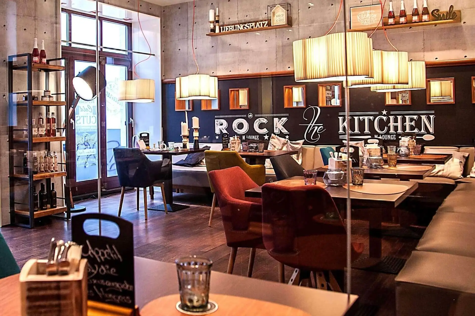 Ein gemütliches, modernes Restaurant mit warmer Beleuchtung, bunten Stühlen, Holztischen, Wanddekorationen und einem Schild mit der Aufschrift "Rock the Kitchen". Flaschen und Dekoration sind auf Regalen und Tischen ausgestellt.