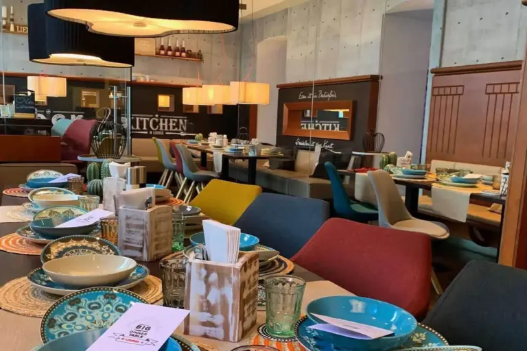 Ein modernes Restaurant mit bunten Stühlen, gedeckten Tischen mit gemusterten Tellern, Gläsern, Servietten und Speisekarten. Warme Beleuchtung und eine gemütliche, zeitgenössische Atmosphäre mit dekorativen Elementen sind im ganzen Raum zu sehen.