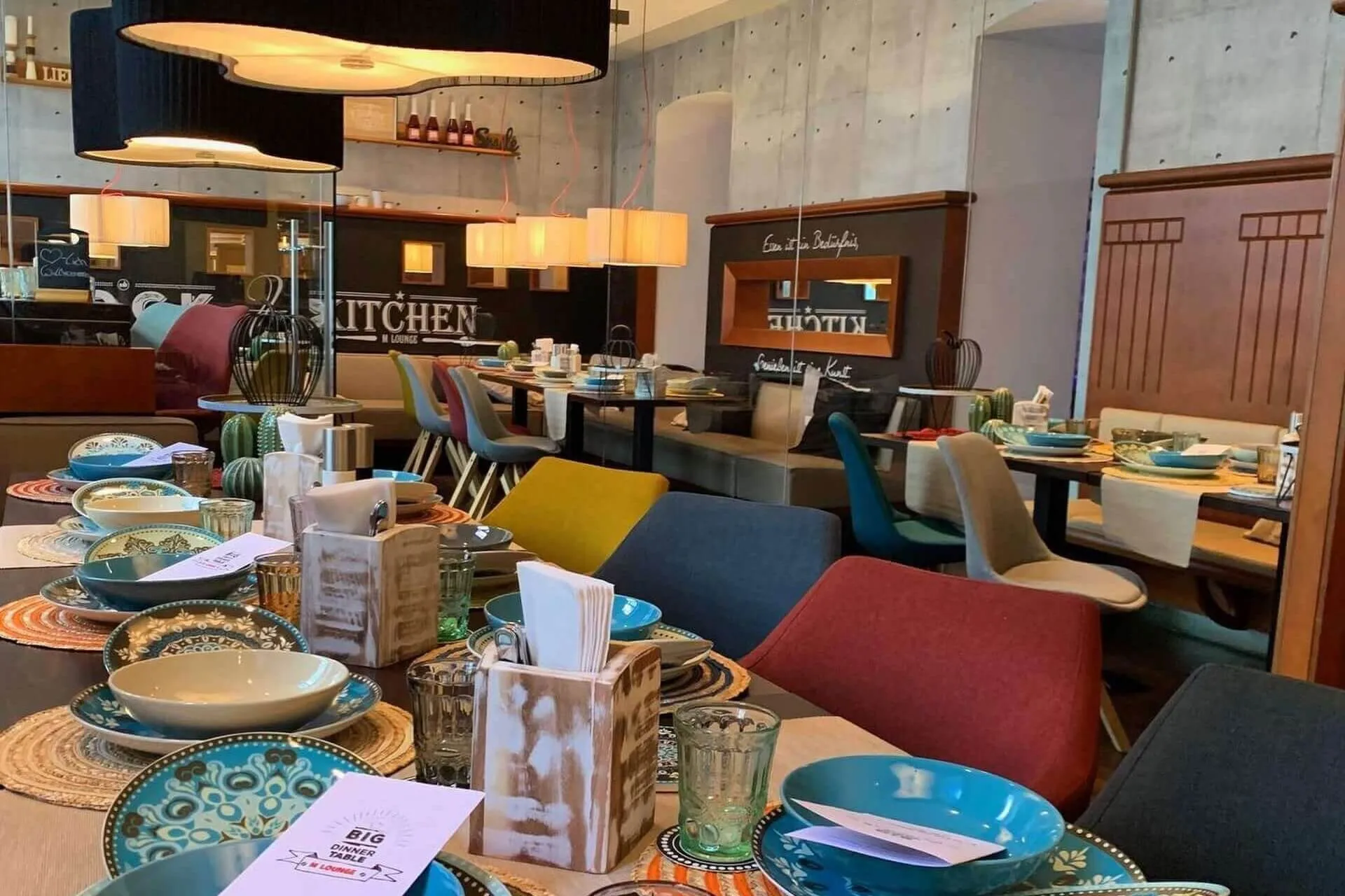 Ein modernes Restaurant mit bunten Stühlen, gedeckten Tischen mit gemusterten Tellern, Gläsern, Servietten und Speisekarten. Warme Beleuchtung und eine gemütliche, zeitgenössische Atmosphäre mit dekorativen Elementen sind im ganzen Raum zu sehen.