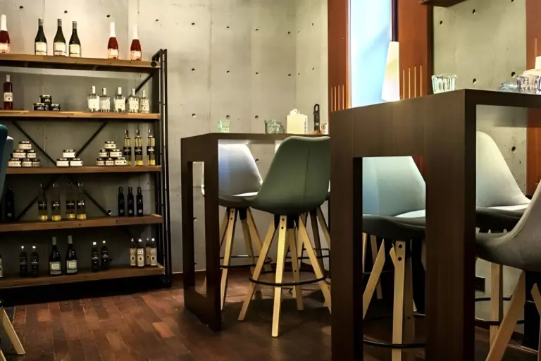 Moderne Bar mit Stehtischen und grün gepolsterten Hockern, Holzregalen mit Flaschen und Gläsern und einer Betonwand im Hintergrund. Die warme Beleuchtung schafft eine gemütliche, einladende Atmosphäre.
