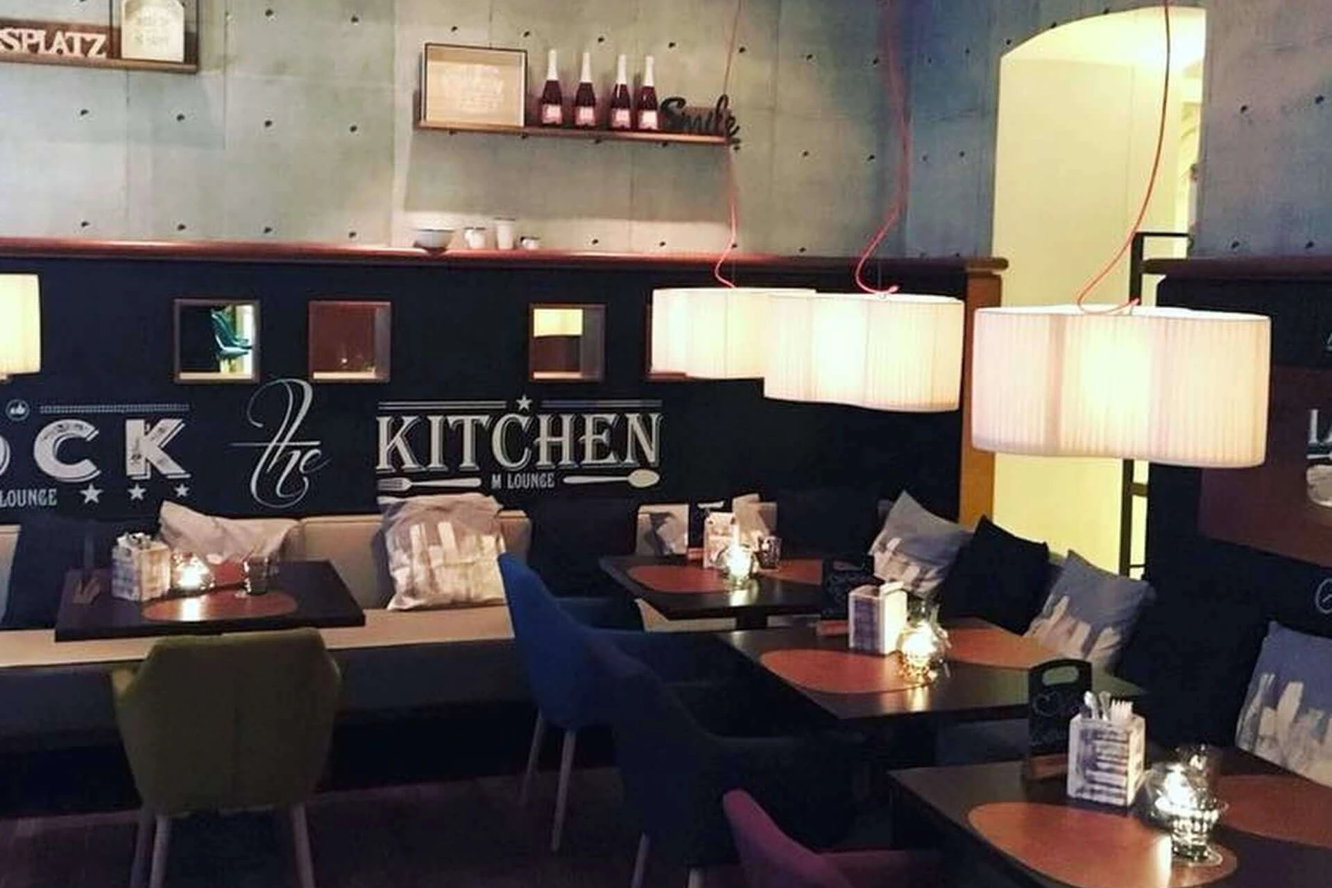 Gemütliches Restaurantinterieur mit gepolsterten Sitzgelegenheiten, kleinen Tischen, hängenden Hängelampen und dekorativer Wandmalerei, die "The Kitchen" zeigt. Regale mit Flaschen und sanfte Beleuchtung schaffen eine warme, einladende Atmosphäre.