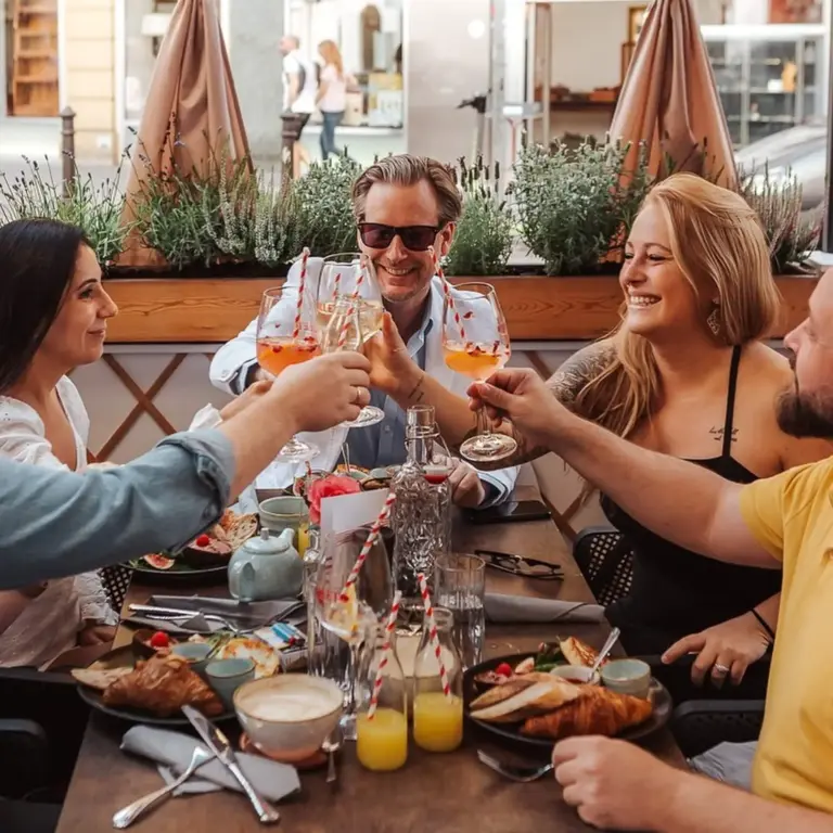 Vier Personen sitzen an einem Tisch in einem Restaurant im Freien, lächeln und erheben ihre Gläser zum Anstoßen. Auf dem Tisch stehen Teller mit Essen, Saft und Kaffeetassen. Grüne Pflanzen säumen den Hintergrund.