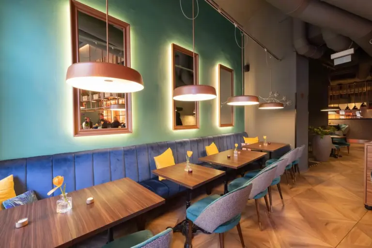 Modernes Café-Interieur mit tealfarbenen Wänden, Holztischen, blau gepolsterten Sitzmöbeln und gelben Akzentkissen. Große runde Hängelampen hängen darüber, und Spiegel schmücken die Wand und reflektieren den gemütlichen Raum.