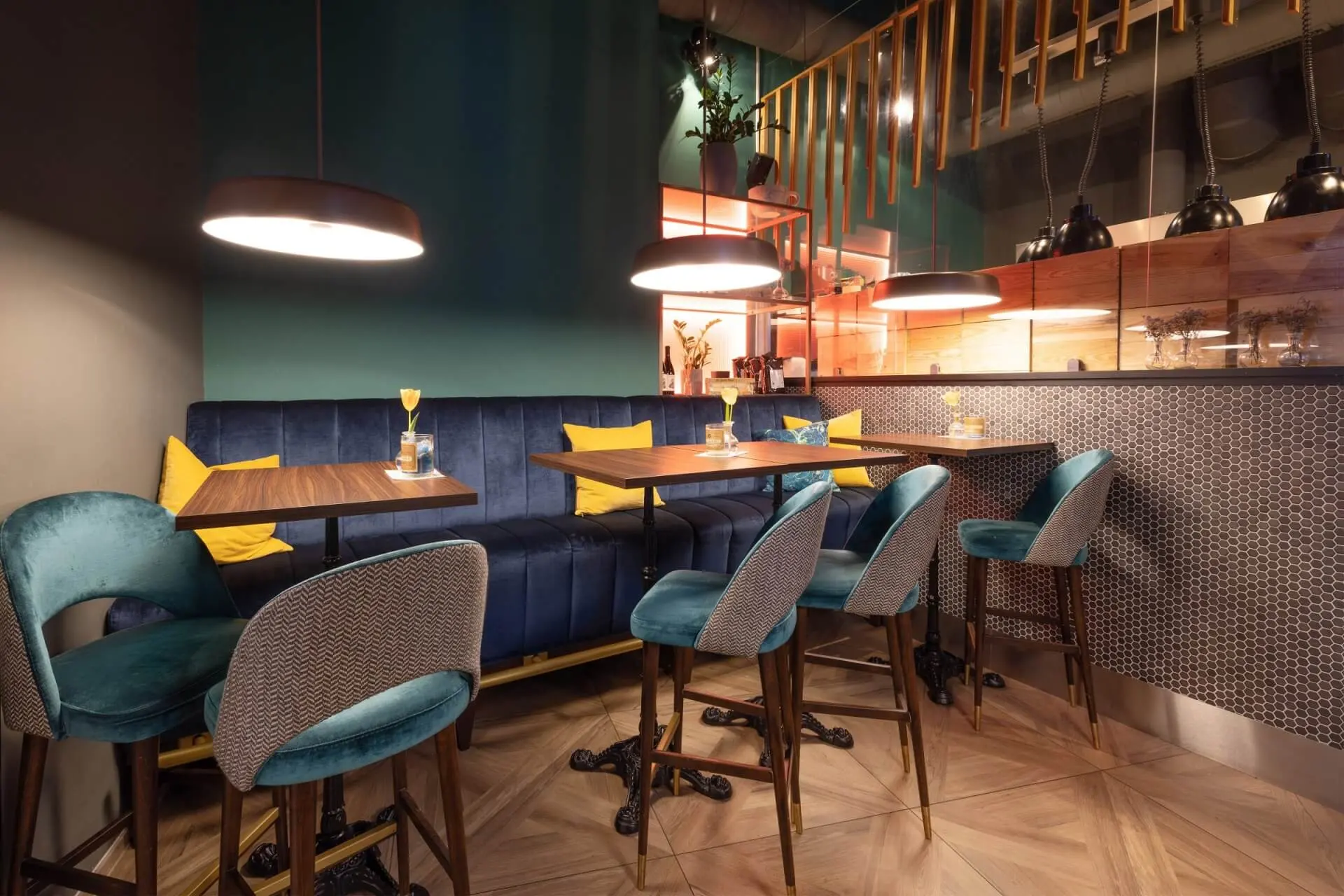 Ein modernes, stilvolles Café-Interieur mit blaugrünen und blauen gepolsterten Stühlen, Holztischen, Hängeleuchten und einer blauen gepolsterten Bank an der Wand. Der Raum hat eine warme, einladende Beleuchtung und eine moderne Einrichtung.