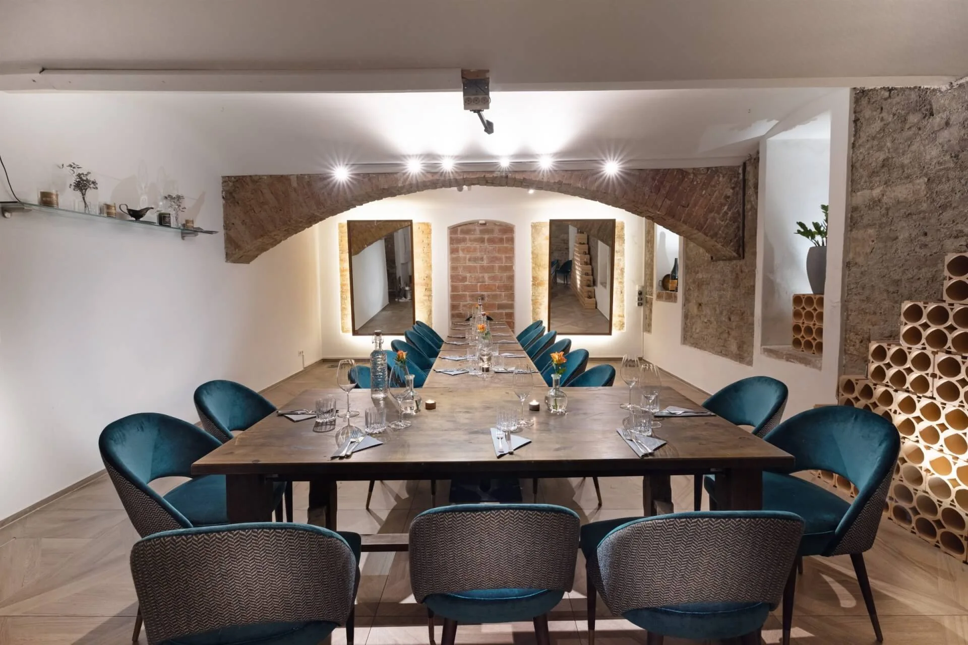 Ein modernes Esszimmer mit einem Holztisch für acht Personen, umgeben von tealfarbenen Samtstühlen. Der Raum verfügt über freiliegende Ziegelbögen, dekorative Wandausschnitte und eine warme Raumbeleuchtung.