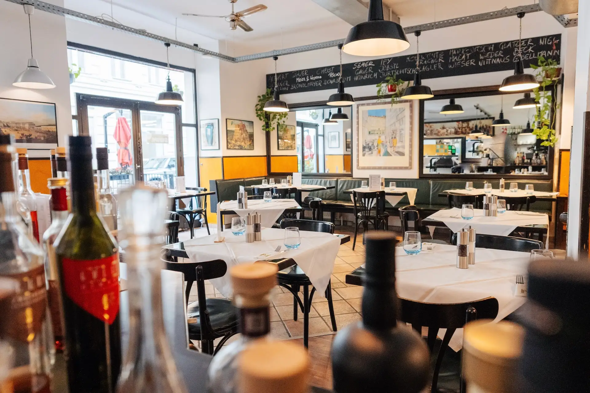 Ein helles, gemütliches Restaurant mit Holzstühlen, weißen Tischdecken, Flaschen im Vordergrund, großen Fenstern, Wandbildern, hängenden Pflanzen und einer Speisekarte auf einer Kreidetafel an der Wand. Der Raum ist leer und strahlt eine einladende Atmosphäre aus.