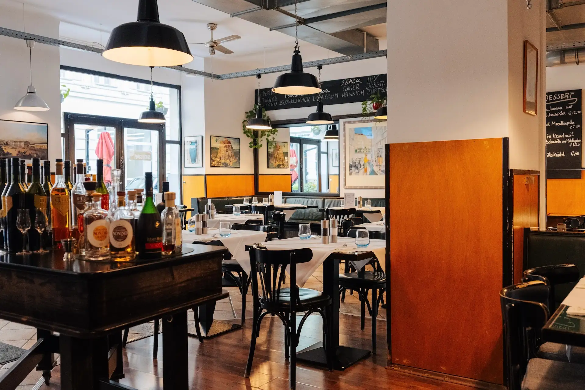 Ein gemütliches, helles Restaurant mit Holzböden, schwarzen Stühlen und für die Gäste gedeckten Tischen. Auf einem Servierwagen stehen Wein- und Spirituosenflaschen. Große Fenster lassen Tageslicht herein, und gerahmte Kunstwerke schmücken die Wände.