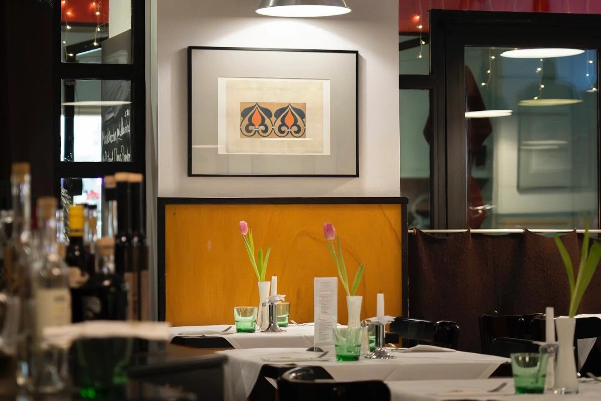 Eine gemütliche Restaurantatmosphäre mit ordentlich gedeckten Tischen, weißen Tischdecken, grünen Gläsern und einzelnen rosa Tulpen in Vasen. An einer mit Holz vertäfelten Wand hängen unter sanfter Beleuchtung Kunstwerke mit zwei flammenartigen Formen.