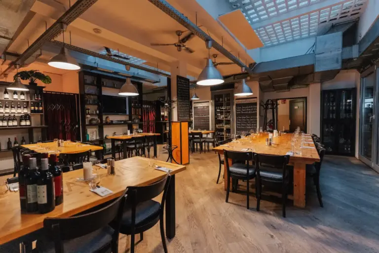 Magazin Ein gemütliches, modernes Restaurant mit Holztischen, schwarzen Stühlen, Weinflaschen in Regalen, Speisekarten auf Kreidetafeln, Pendelleuchten und einer Decke mit Oberlicht. Die Tische sind gedeckt, und der Raum wirkt bereit für die Gäste.