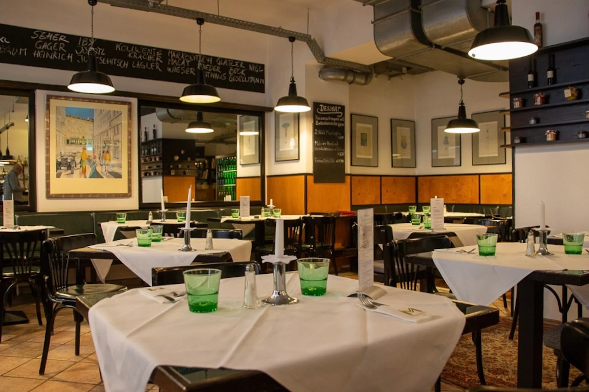 Ein gemütliches Restaurant mit ordentlich gedeckten Tischen, grünen Gläsern, Speisekarten und Kerzen. An den Wänden hängen gerahmte Kunstwerke, und darüber ist eine Kreidetafel mit der Speisekarte angebracht. Schwarze Stühle und eine warme Beleuchtung sorgen für eine einladende Atmosphäre.