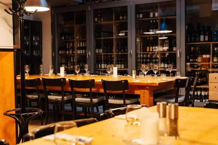 Ein gemütliches Restaurantinterieur mit Holztischen, auf denen Weingläser und Besteck stehen. An der Rückwand reihen sich Regale voller Weinflaschen aneinander, und schwarze Stühle umgeben die Tische. Warme Beleuchtung sorgt für eine einladende Atmosphäre.