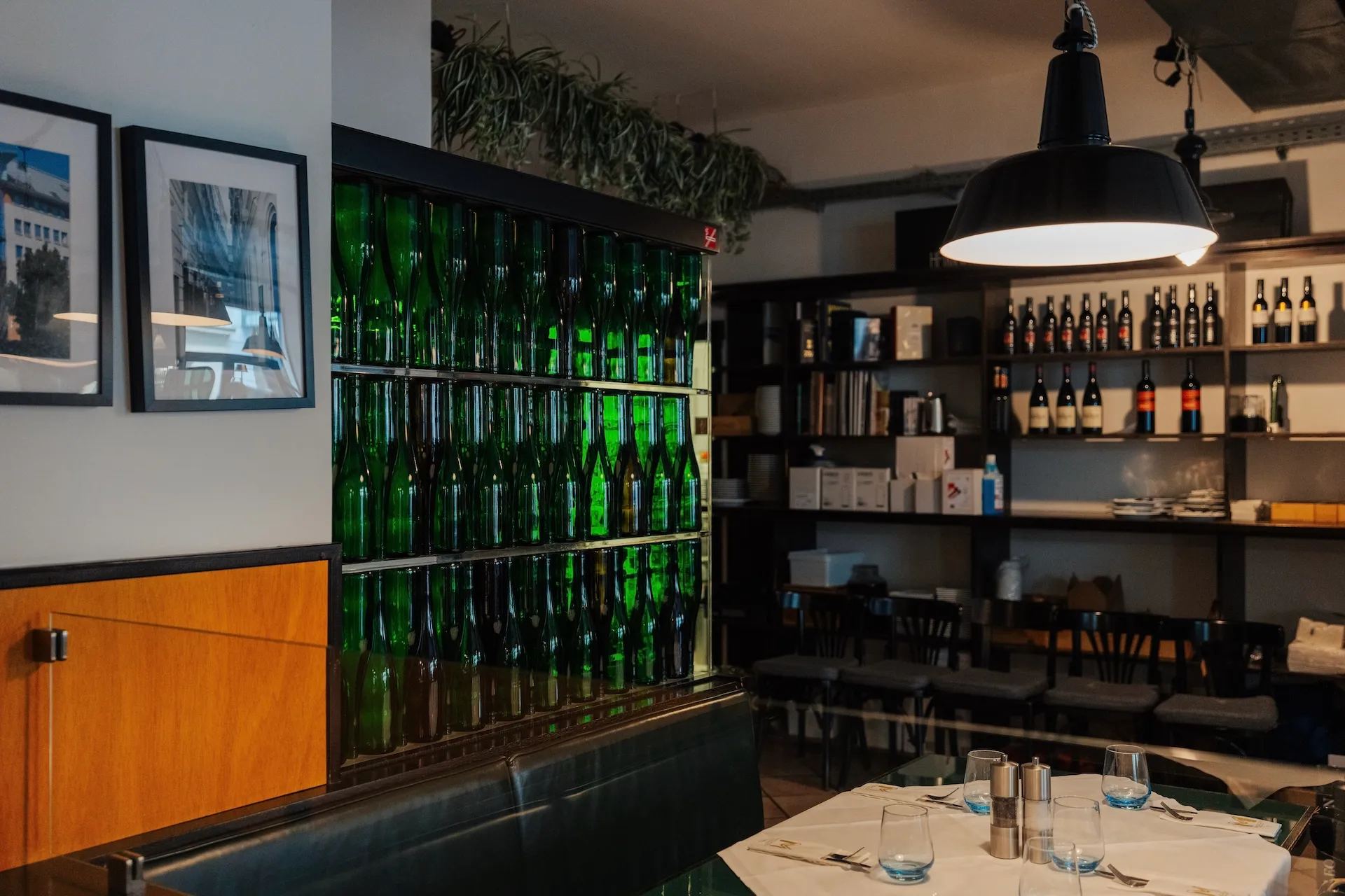 Ein gemütliches Restaurantinterieur mit grünen Flaschen, die in einer Vitrine gestapelt sind, Weinregalen, gerahmten Fotos an der Wand, einem gedeckten Esstisch mit Gläsern und einer großen schwarzen Hängelampe über dem Tisch.