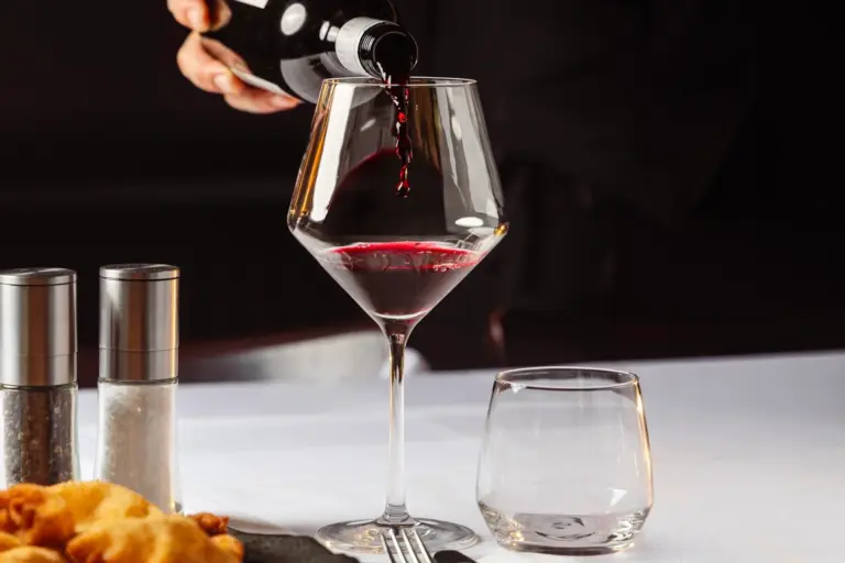 Eine Person schenkt Rotwein in ein Glas auf einer weißen Tischdecke ein; daneben sind ein Wasserglas, Salz- und Pfefferstreuer sowie Speisen zu sehen.
