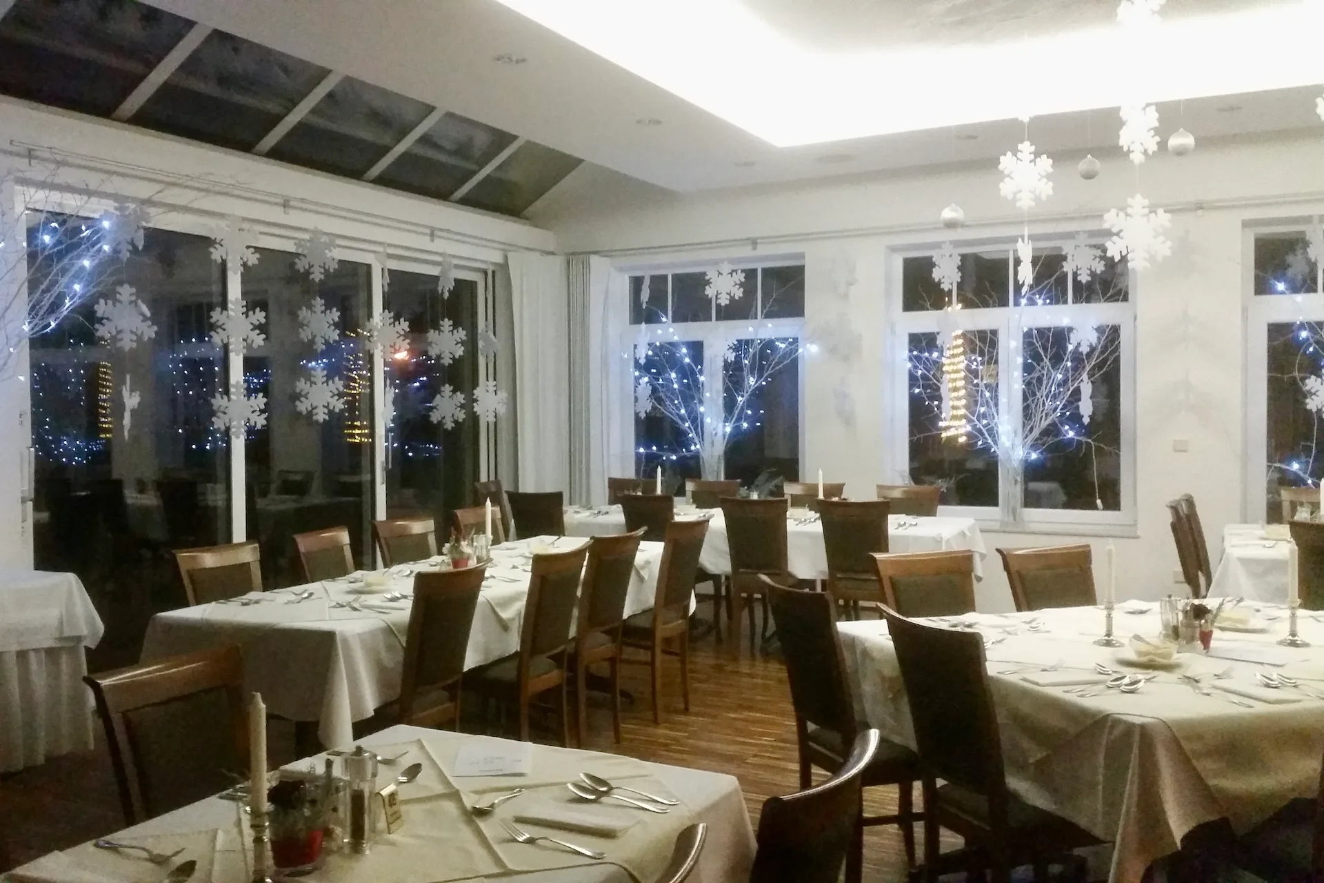 Ein Esszimmer, das für das Abendessen hergerichtet ist, mit Tischen, die mit weißen Tischdecken bedeckt sind, und an den Fenstern mit hängenden Schneeflockenausschnitten und beleuchteten Zweigarrangements geschmückt.