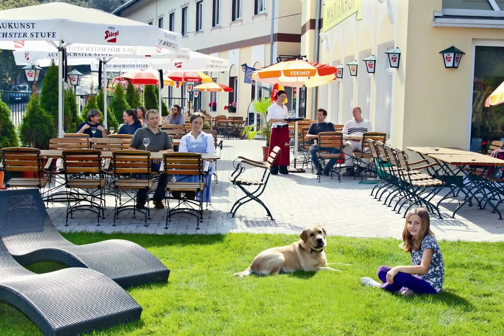 Ein Mädchen sitzt auf dem Rasen neben einem großen Hund vor der Terrasse eines Restaurants im Freien, auf der Gäste sitzen und Sonnenschirme stehen.