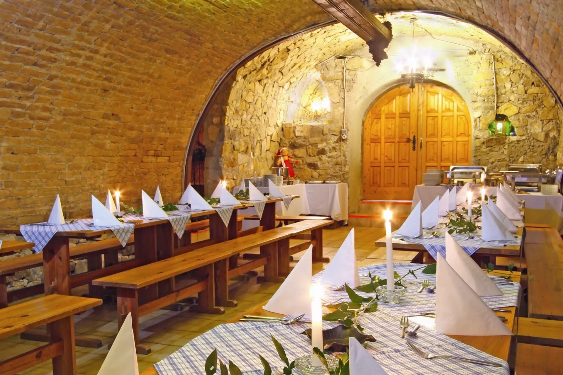 Lange Holztische, gedeckt für ein Essen in einem Keller mit Steinwänden, Gewölbedecken und brennenden Kerzen.