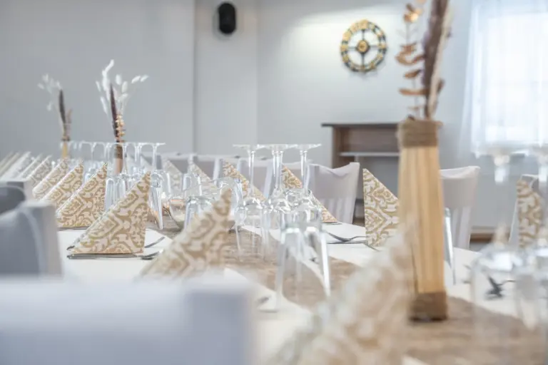 Elegant gedeckter Tisch mit gemusterten gefalteten Servietten, umgedrehten Weingläsern und schlichter Tischdekoration in einem hellen, sanft beleuchteten Raum. Im Hintergrund sind eine Wanduhr und ein Konsolentisch zu sehen.