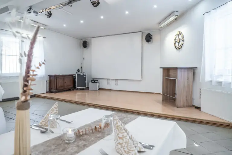 Eine kleine Bühne mit einem Podium, einem großen Bildschirm und einer Audioanlage in einem hellen, modernen Raum. Im Vordergrund ist ein Esstisch mit gefalteten Servietten, Besteck und einer Kerze gedeckt.