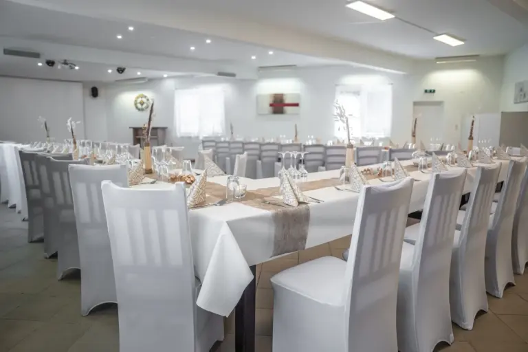Ein geräumiger Bankettsaal mit langen, mit weißen Tischdecken bedeckten Tischen und ordentlich aufgestellten Stühlen, die für eine Veranstaltung mit gefalteten Servietten, Gläsern und schlichten beigen und weißen Tafelaufsätzen gedeckt sind. Natürliches Licht füllt den Raum.