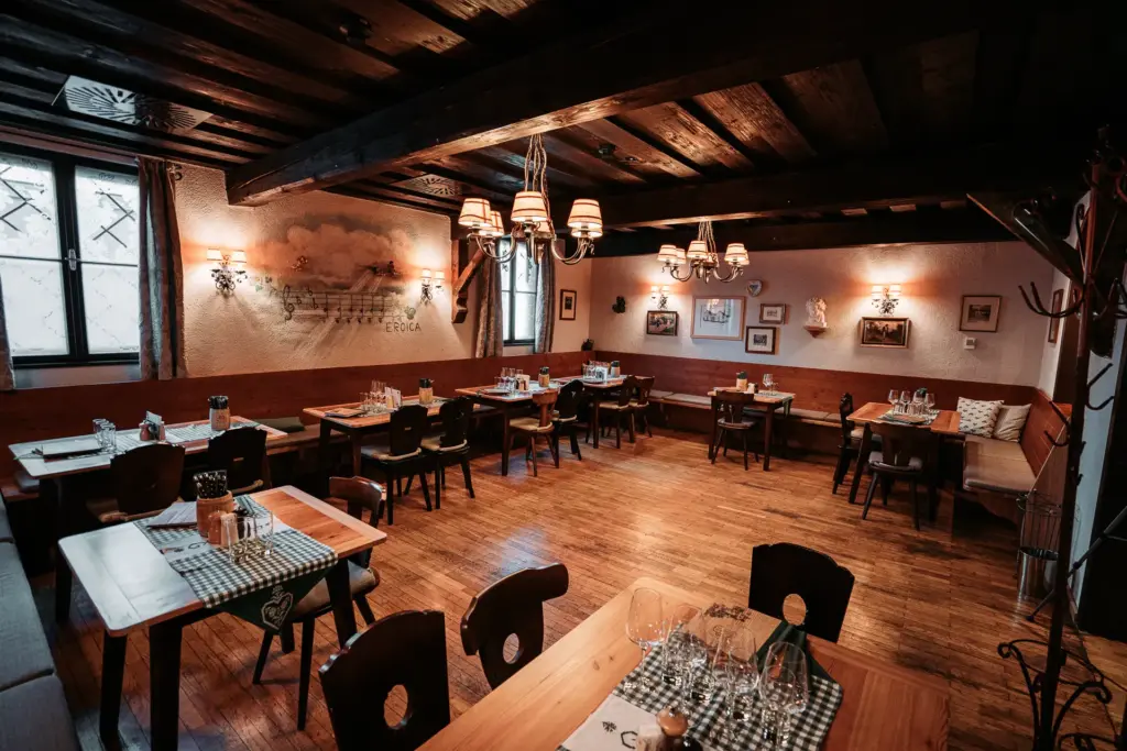 Ein gemütlicher, rustikaler Speisesaal mit Holzböden und -balken, mit karierten Servietten gedeckten Tischen, Glaswaren und sanfter Beleuchtung durch Kronleuchter. Gerahmte Bilder und Wandkunst schmücken die warm getönten Wände.