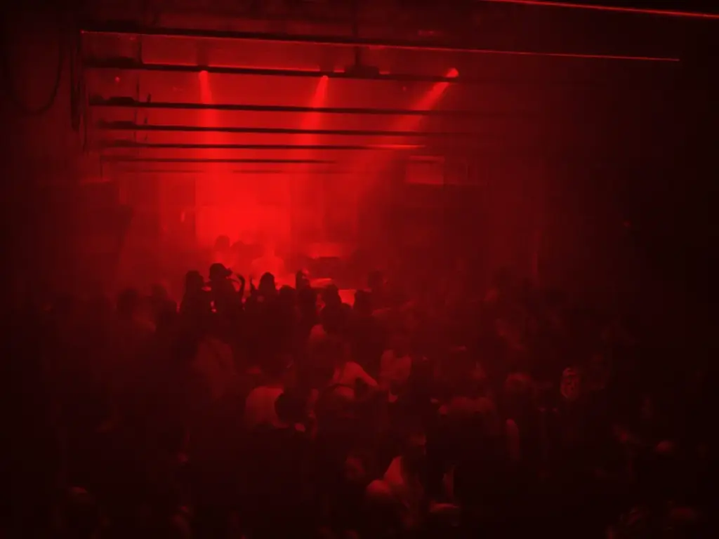 Eine große Menschenmenge in einem dunklen, überdachten Raum im Club Praterstraße, beleuchtet von intensivem rotem Licht und Strahlen, die eine rauchige, energiegeladene Atmosphäre schaffen, die typisch für einen Nachtclub oder ein Konzert ist.