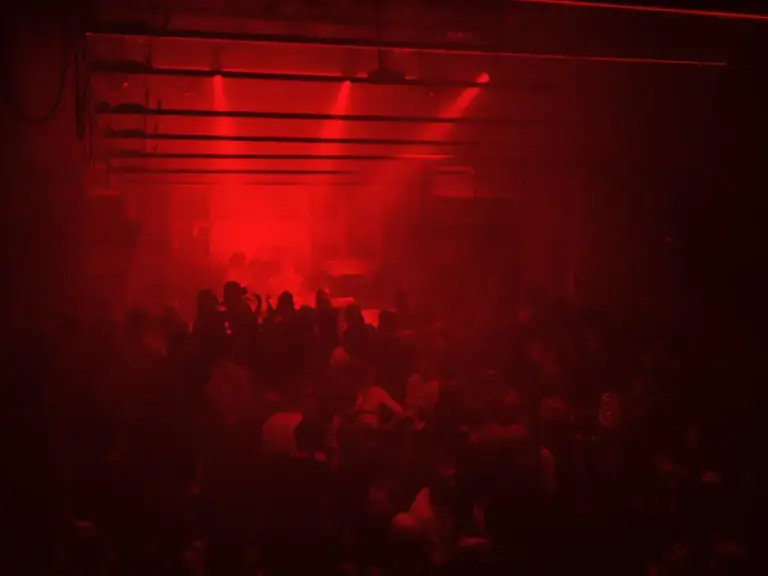 Eine große Menschenmenge in einem dunklen, überdachten Raum im Club Praterstraße, beleuchtet von intensivem rotem Licht und Strahlen, die eine rauchige, energiegeladene Atmosphäre schaffen, die typisch für einen Nachtclub oder ein Konzert ist.