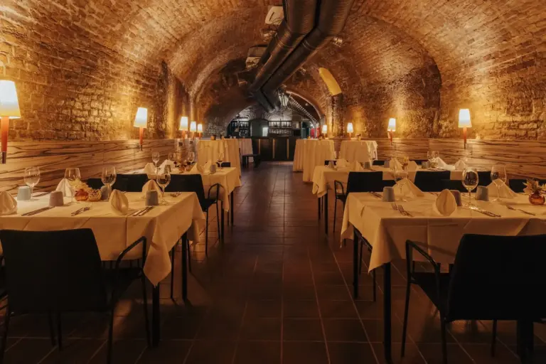 Mercure Grand Hotel Biedermeier Ein gemütliches Restaurant mit Gewölbedecken aus Ziegeln, die von Wandlampen warm beleuchtet werden. Die Tische sind mit weißen Tüchern, Gläsern und Servietten gedeckt, was eine einladende, elegante Atmosphäre schafft.