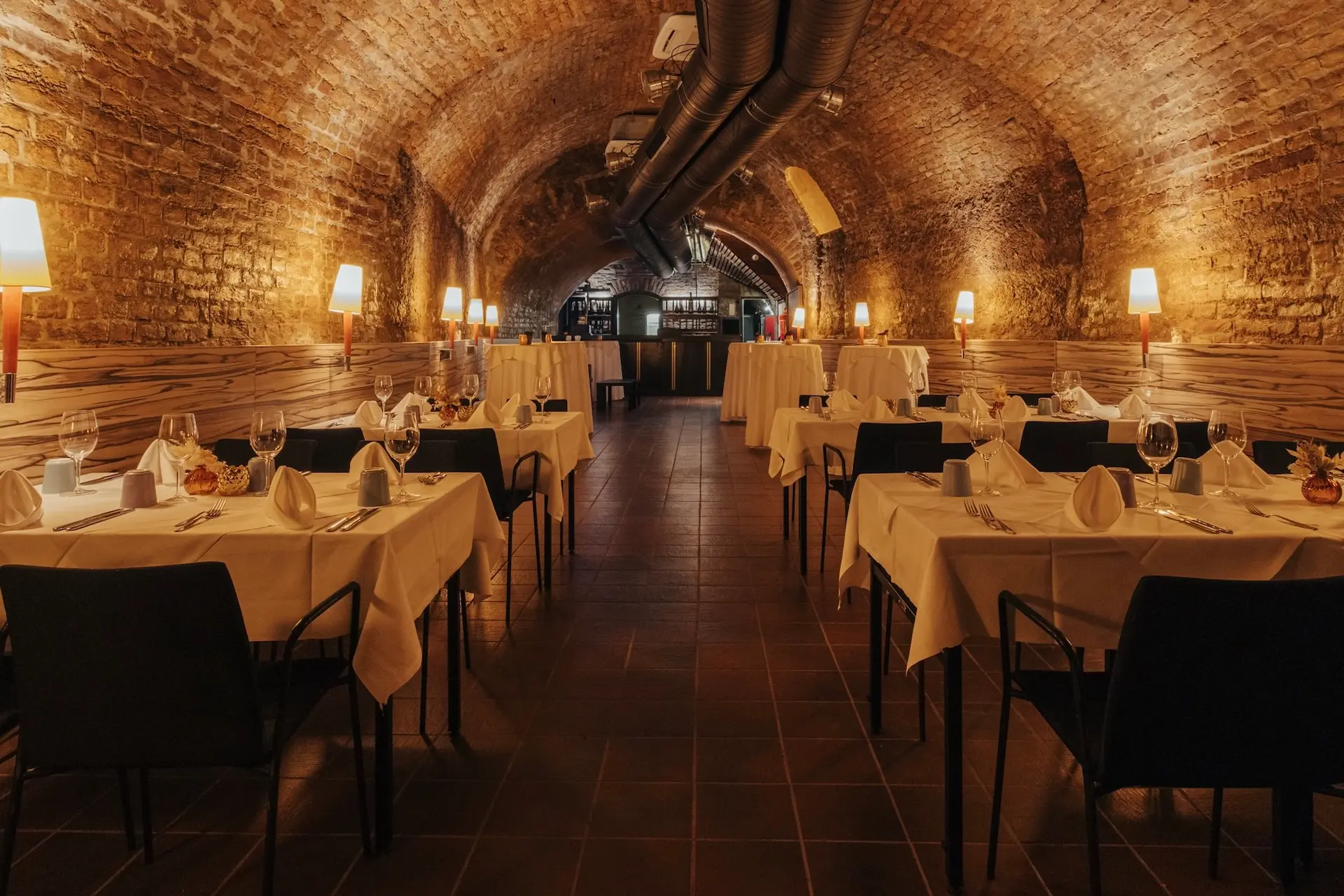 Mercure Grand Hotel Biedermeier Ein gemütliches Restaurant mit Gewölbedecken aus Ziegeln, die von Wandlampen warm beleuchtet werden. Die Tische sind mit weißen Tüchern, Gläsern und Servietten gedeckt, was eine einladende, elegante Atmosphäre schafft.