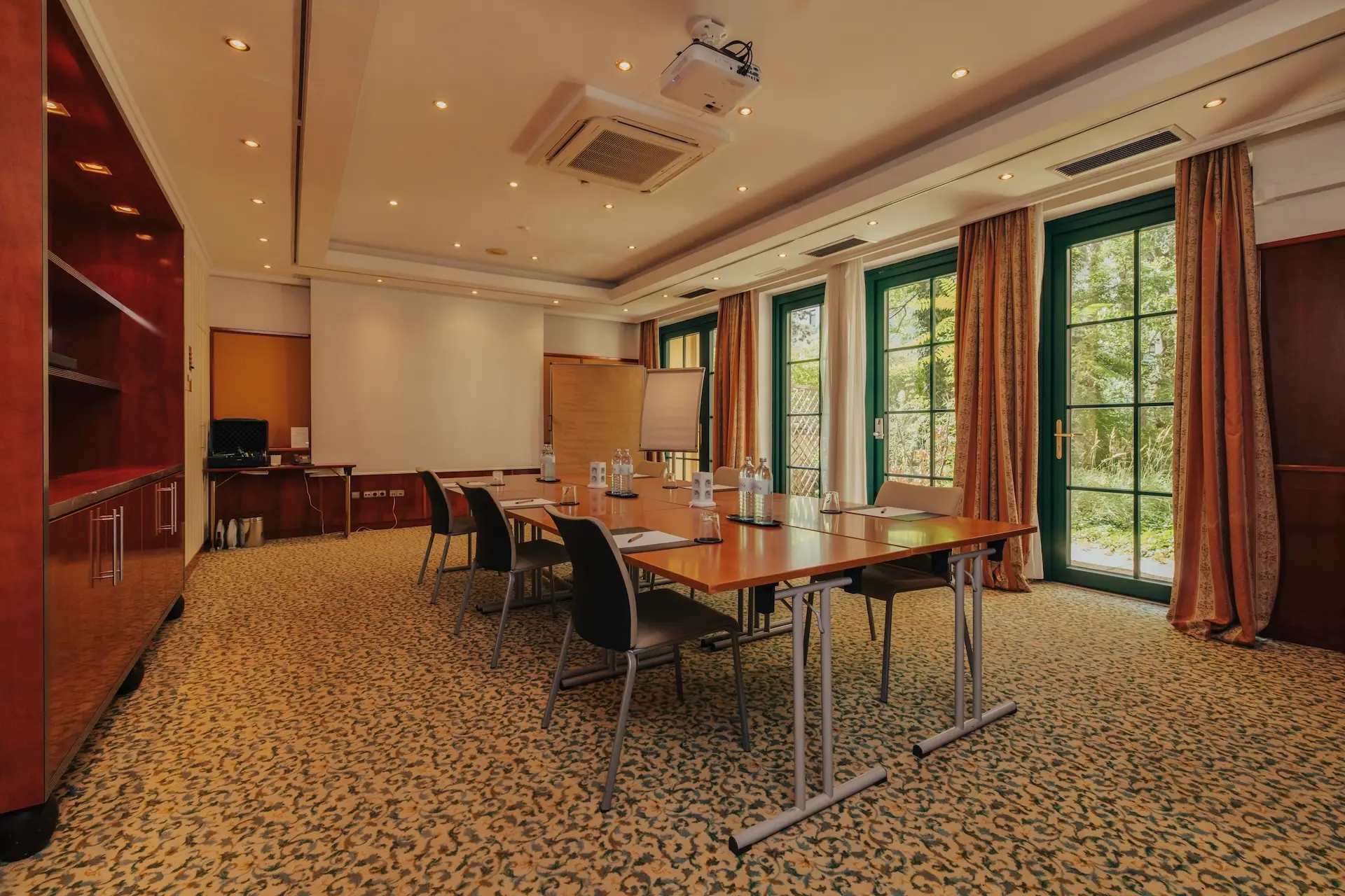 Ein gut beleuchteter Konferenzraum mit großen Fenstern, einem langen Tisch mit Wasserflaschen und Notizblöcken, schwarzen Stühlen, einem Flipchart und eingebauten Holzregalen an der Wand.