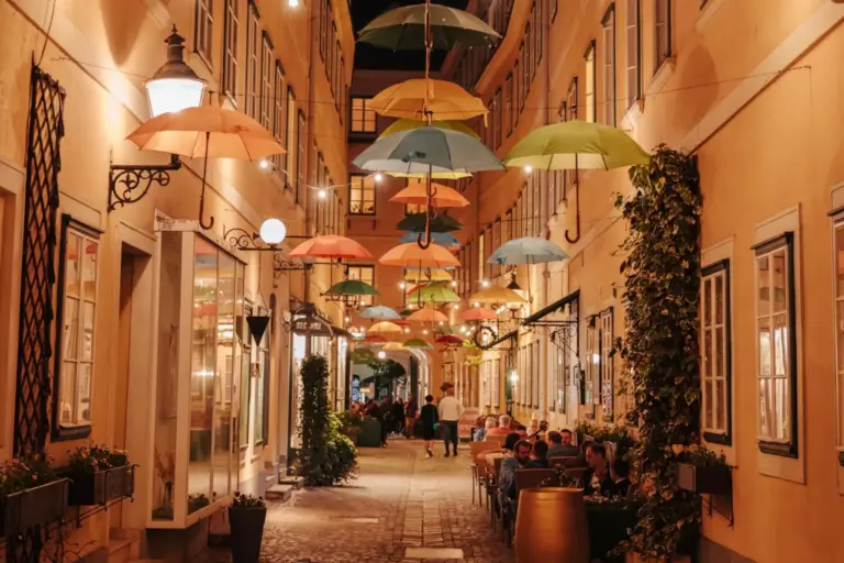 In einer engen, warm beleuchteten Gasse hängen nachts bunte Sonnenschirme über dem Kopf. Entlang des kopfsteingepflasterten Weges sitzen Menschen an Cafétischen im Freien, umgeben von Pflanzen und hohen Gebäuden mit vielen Fenstern.