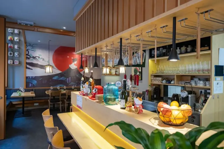Ein modernes japanisches Restaurant mit Holzmöbeln, einer Theke mit Getränken und Obst, hängenden Gläsern und einem Wandgemälde mit einer Geisha und einer roten Sonne im Hintergrund. Im Hintergrund sind Tische und Kabinen zu sehen.