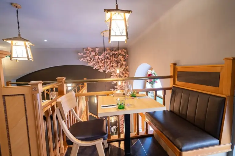 Ein gemütlicher Sitzbereich im Restaurant mit einem Holztisch, einer gepolsterten Bank und einem Stuhl. Eine dekorative Beleuchtung hängt von der Decke, und rosa Kirschblütenzweige verleihen dem Hintergrund Charme. Gläser und Servietten stehen auf dem Tisch.