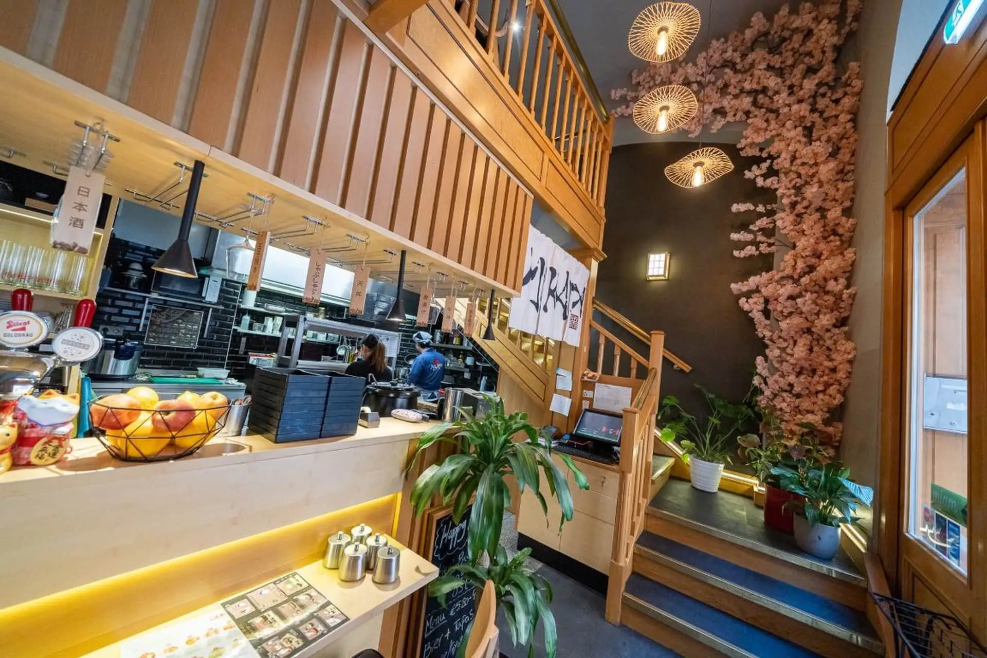 Modernes japanisches Restaurant mit Holzdekor, hängenden Laternen, einer Treppe, Topfpflanzen und einer mit rosa Kirschblüten geschmückten Wand. Die Küche ist sichtbar und die Atmosphäre ist hell und einladend.