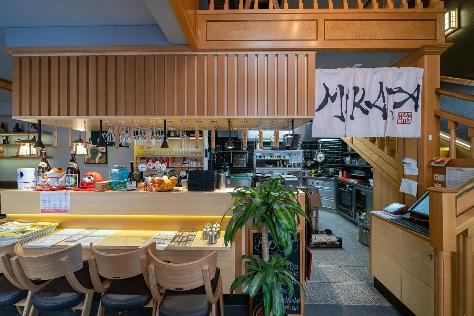 Ein gemütliches japanisches Restaurant mit Holzstühlen an einem Tresen, einer offenen Küche im Hintergrund und einem weißen Banner mit japanischen Schriftzeichen darüber. Eine Topfpflanze steht neben dem Eingang.
