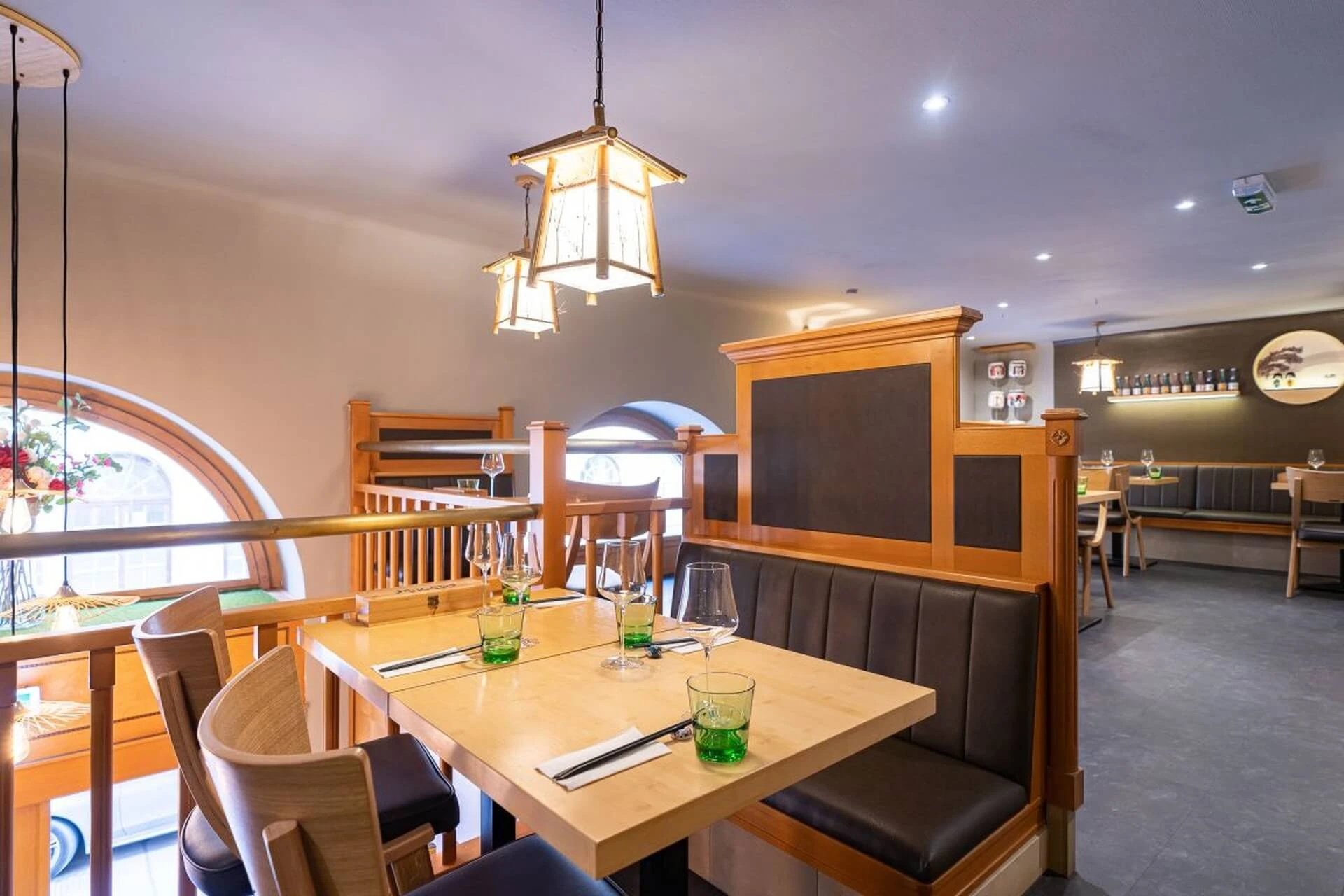 Ein modernes Restaurant mit Holztischen und -stühlen, braunen gepolsterten Sitzgelegenheiten, Hängelaternen und Gedecken mit grünen Gläsern. Natürliches Licht fällt durch gewölbte Fenster ein.