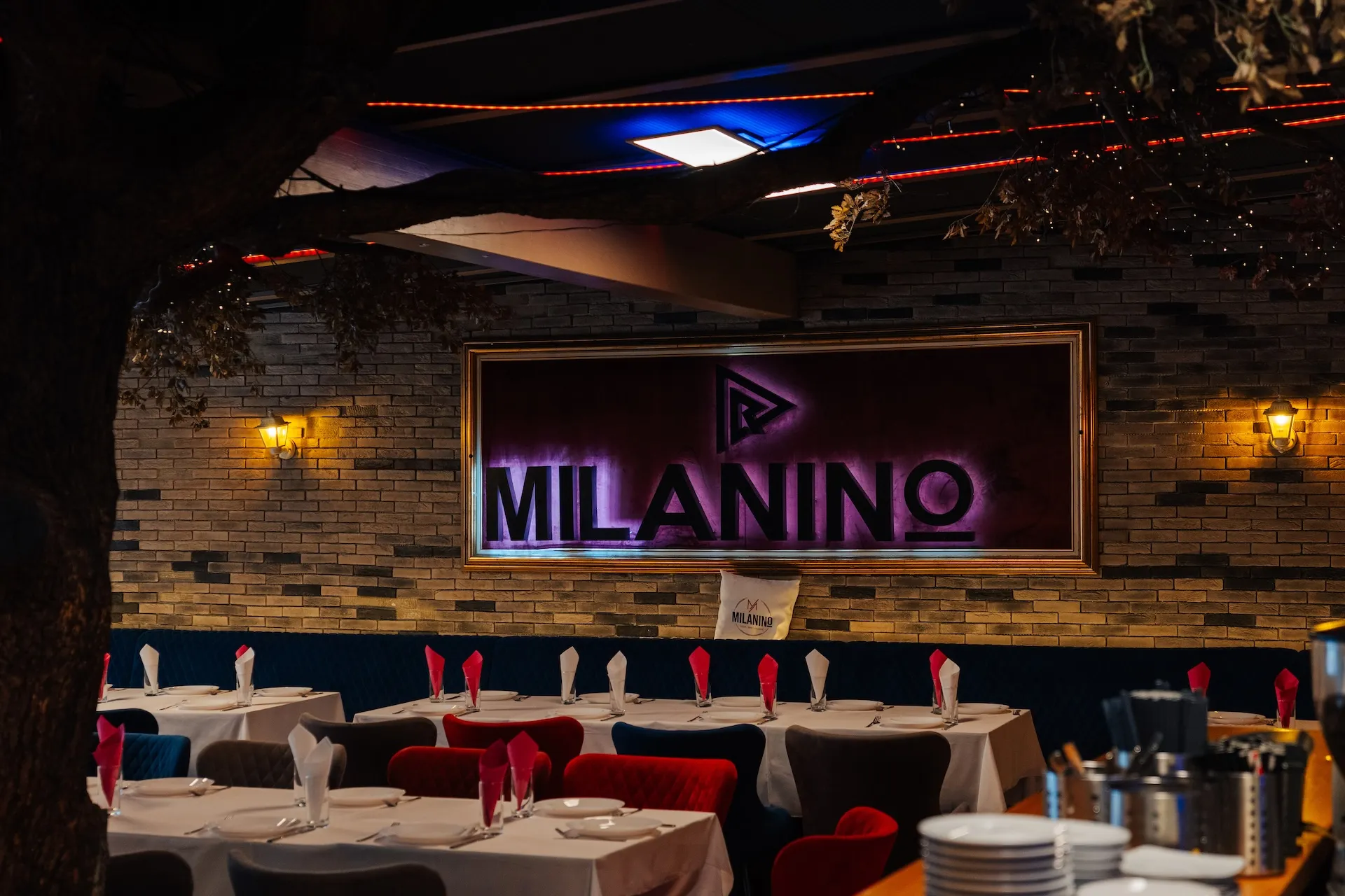 Der Innenraum eines Restaurants mit Tischen, die mit weißen Tischdecken und rot-weißen Servietten gedeckt sind. Auf der Backsteinwand ist ein großes Leuchtschild mit der Aufschrift „MILANINO“ zu sehen. Warme Beleuchtung und ein dekorativer Baum sind zu erkennen.