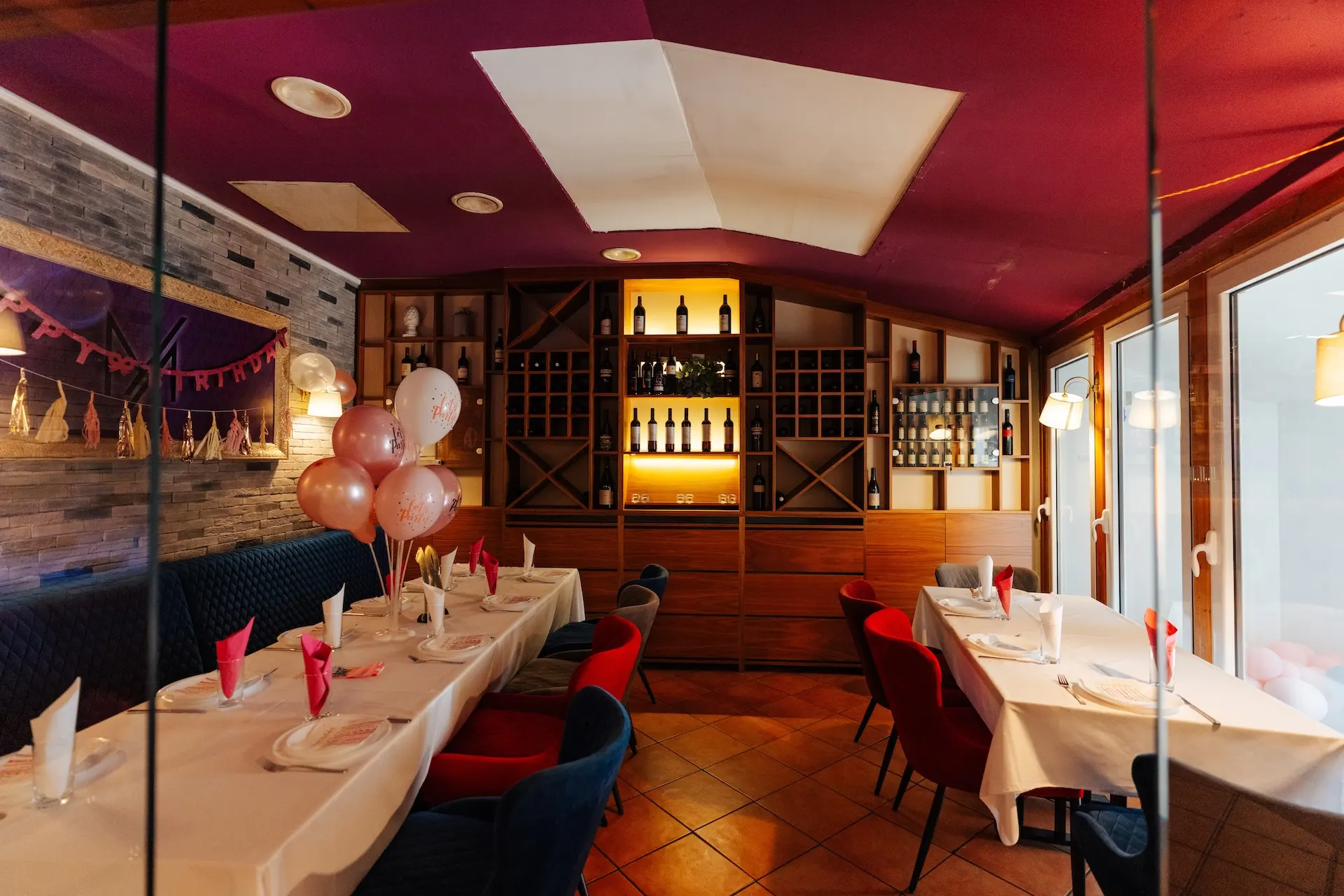 Ein festlich geschmückter Restaurantbereich mit Luftballons, einem Geburtstagsbanner, Tischen, die mit rosa Servietten gedeckt sind, und einem Weinschrank im Hintergrund. Die warme Beleuchtung sorgt für eine festliche und einladende Atmosphäre.