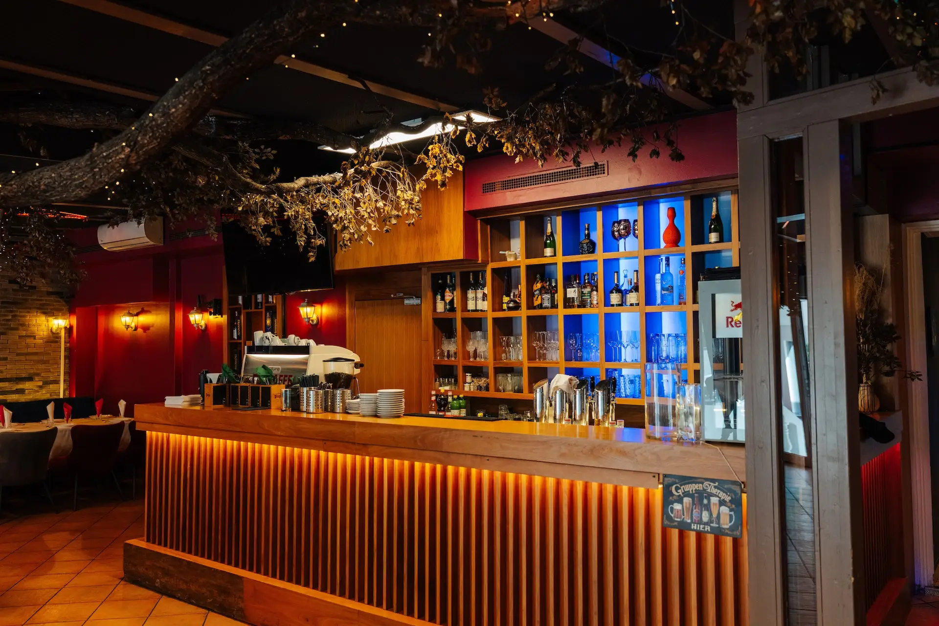 Eine gemütliche Bar mit einer Holztheke, Regalen voller Flaschen und Gläser, warmer Beleuchtung und dekorativen Ästen, die von der Decke hängen.