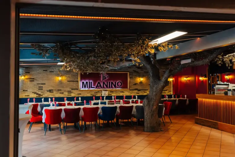 Milanino Ein Restaurantbereich mit roten und blauen Stühlen, die um einen langen Tisch herum stehen, einem künstlichen Baum, warmer Wandbeleuchtung und einem Schild mit der Aufschrift „MILANINO“ an einer Backsteinwand.