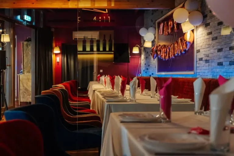 Ein Restaurant mit langen Tischen, die für eine Feier gedeckt sind, mit rot-weißen Servietten, Luftballons und einem „Happy Birthday“-Banner an der Wand. Der Raum ist schwach beleuchtet und mit bunten Stühlen ausgestattet.