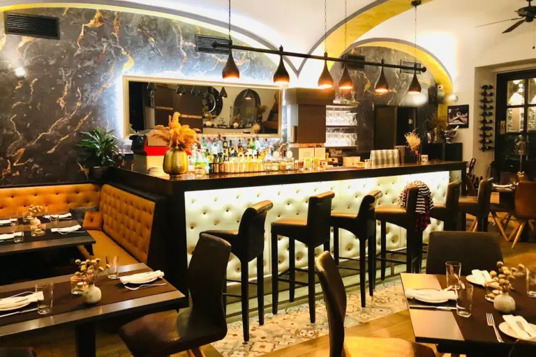 Eine stilvolle Restaurantbar mit getufteten Sitzen, hohen Stühlen, eleganter Beleuchtung und einer Akzentwand aus Marmor. Die Tische sind zum Essen gedeckt, und dekorative Vasen mit Trockenblumen sorgen für einen gemütlichen, modernen Touch.