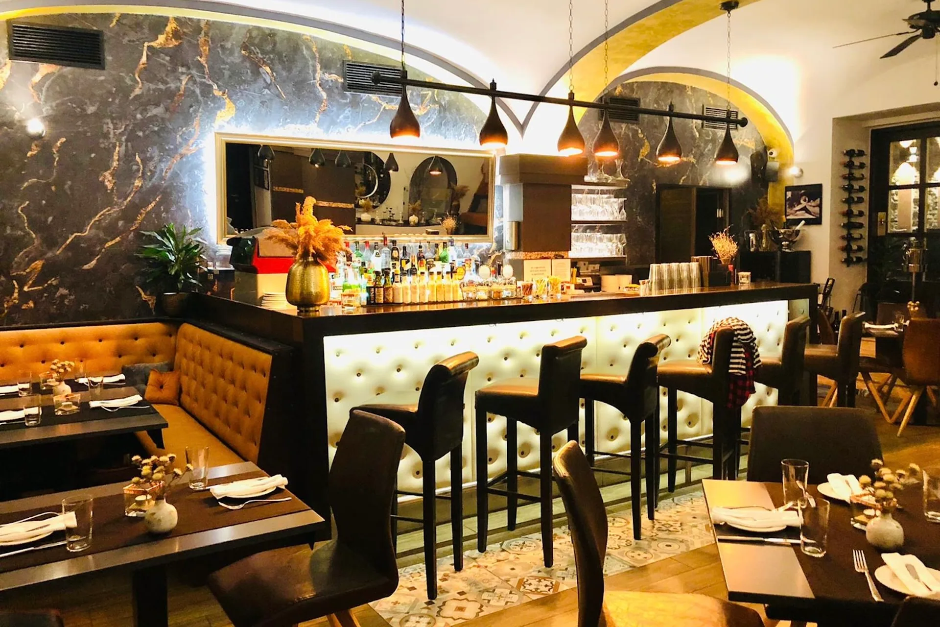 Eine stilvolle Restaurantbar mit getufteten Sitzen, hohen Stühlen, eleganter Beleuchtung und einer Akzentwand aus Marmor. Die Tische sind zum Essen gedeckt, und dekorative Vasen mit Trockenblumen sorgen für einen gemütlichen, modernen Touch.
