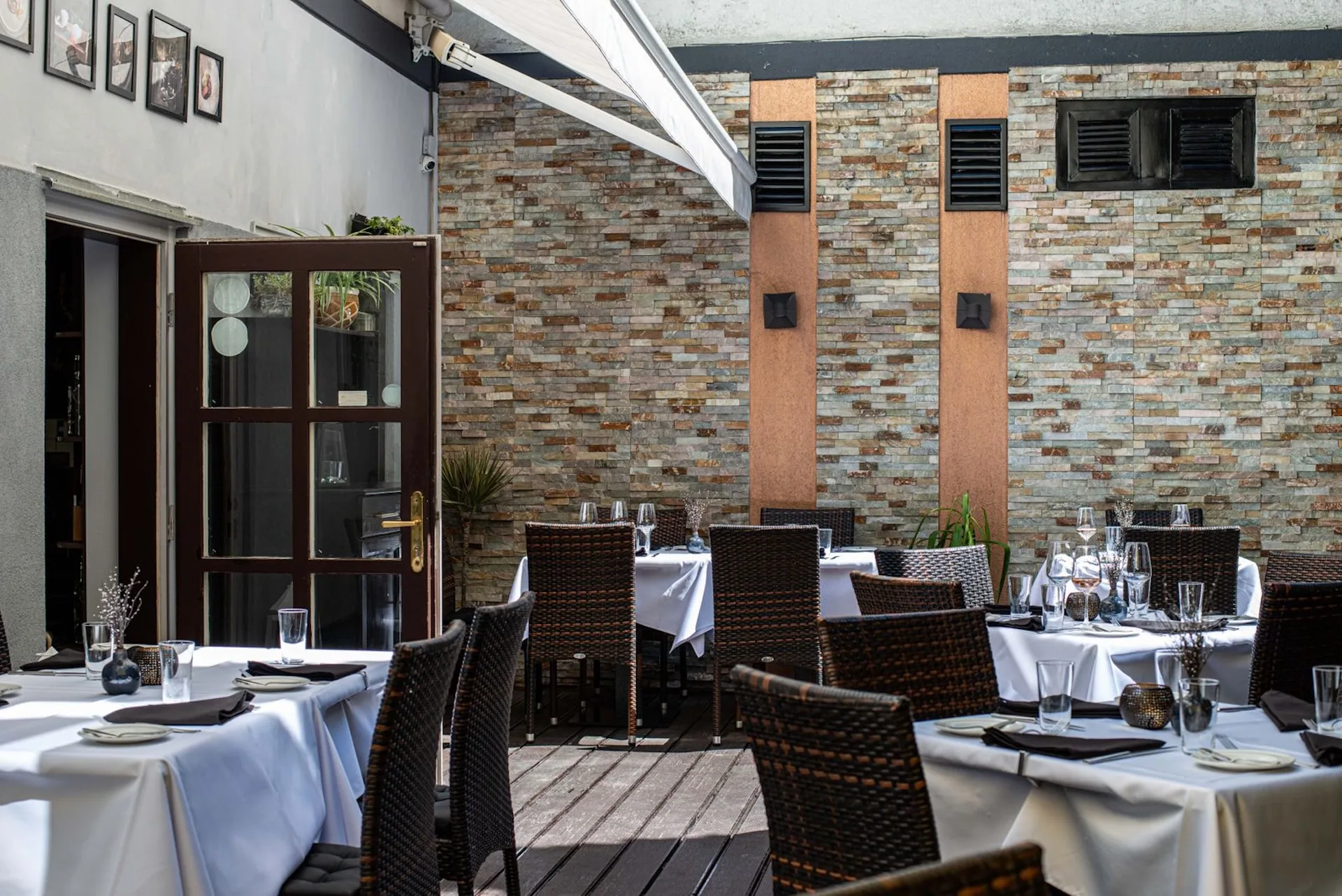 Restaurantterrasse im Freien mit Tischen und Stühlen zum Essen, überdacht von einem Vordach. Die Backsteinwand im Hintergrund, die Glastür auf der linken Seite und das Sonnenlicht erhellen den Bereich.