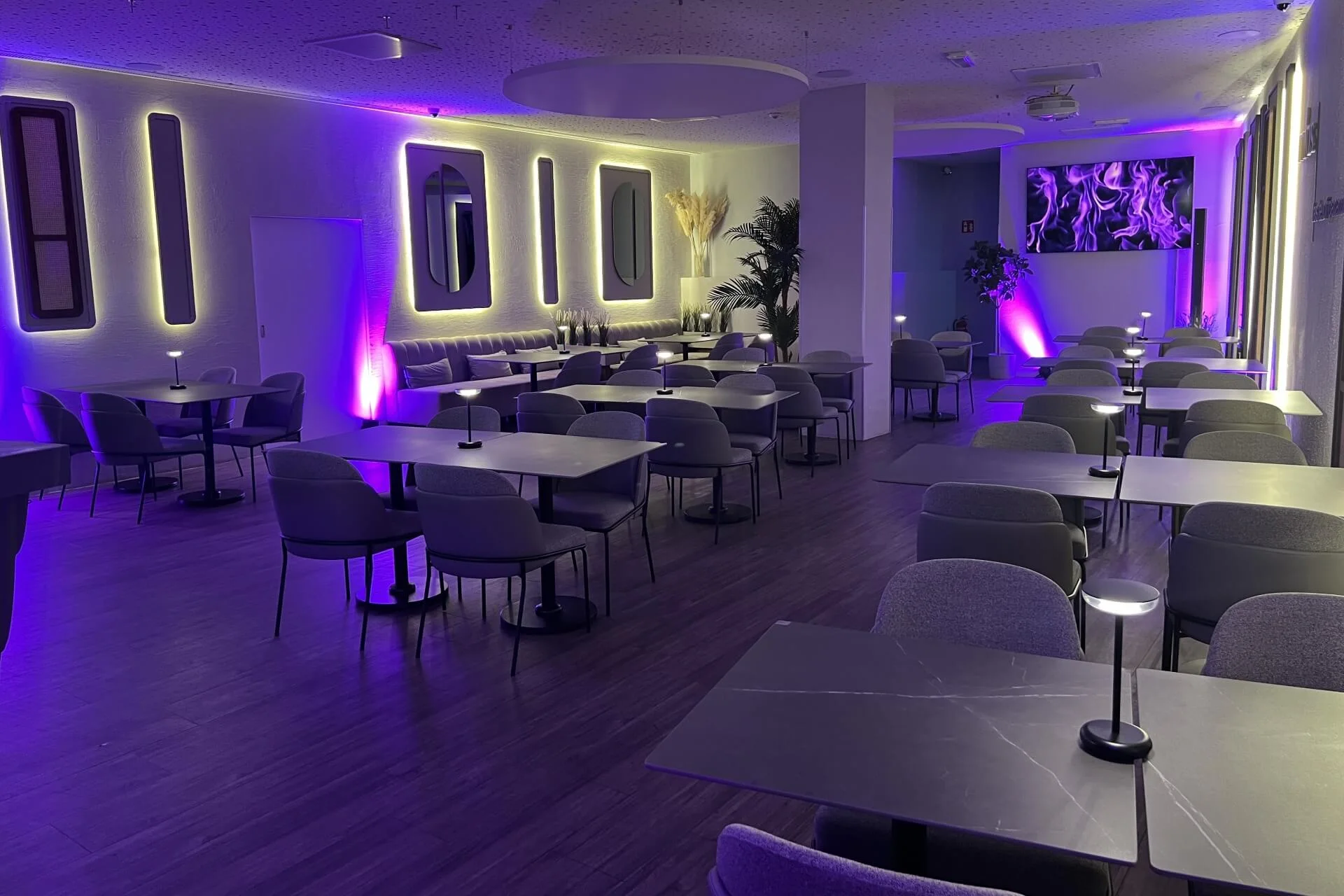 Eine moderne Lounge mit violetter Akzentbeleuchtung, grauen Stühlen, schwarzen Tischen, Wandspiegeln und dekorativen Pflanzen, die eine stilvolle und einladende Atmosphäre schaffen.
